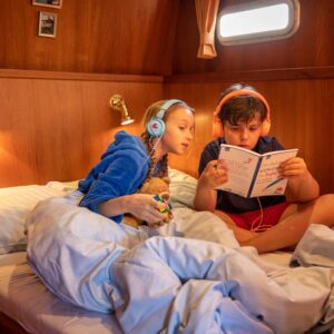 Zwei Kinder teilen sich ein Doppelbett in der Babro Beluga. Sie sitzen auf dem Bett mit Kopfhörern und lesen gemeinsam ein Buch.