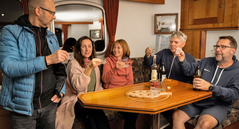 Salon Aquanaut Vintage, 3 Männer und 2 Frauen sitzen am Tisch und stoßen mit kleinen Gläsern an