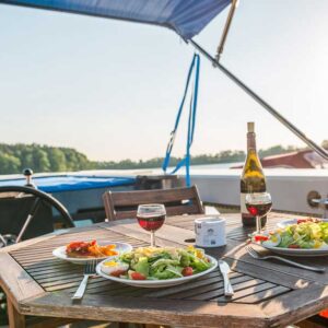 an Deck der Aquino steht ein hübscher Holztisch, gedeckt mit Salat und Wein, darüber ist das Bimini gespannt gegen die Sonne