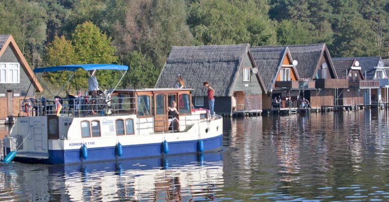 Eine Kormoran fährt an einer reihe Bootshäuser vorbei.