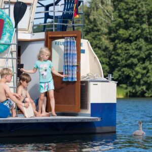 Drei Kinder stehen auf der Badeplattform einer Kormoran. Neben ihnen ist ein Schwan mit zwei Küken.