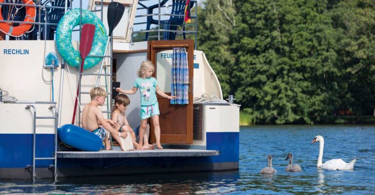 Drei Kinder stehen auf der Badeplattform einer Kormoran. Neben ihnen ist ein Schwan mit zwei Küken.