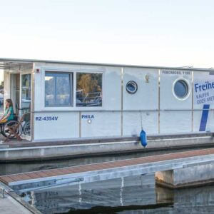 Febo 1180 liegt im Hafen der marine Müritz. Auf der Veranda sitzt eine Rollstuhlfahrerin