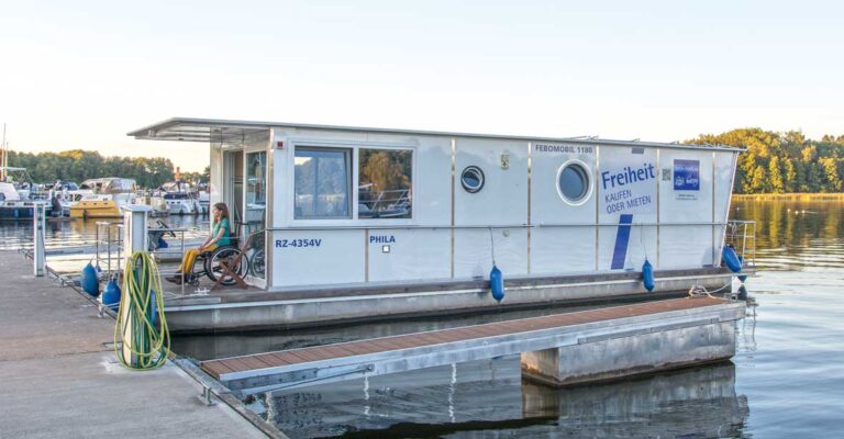 Febo 1180 liegt im Hafen der marine Müritz. Auf der Veranda sitzt eine Rollstuhlfahrerin