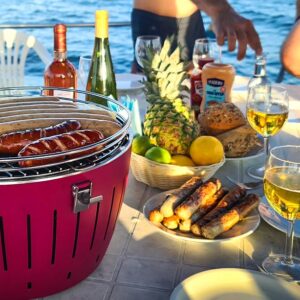 Ein gedeckter Tisch mit einem Grill auf dem bereits einige Würstchen liegen. Daneben ist ein Teller mit bereits Fertigen Würstchen. Dazu ein Paar Gläser Wein, ein Korb mit Obst und ein Paar Brötchen.