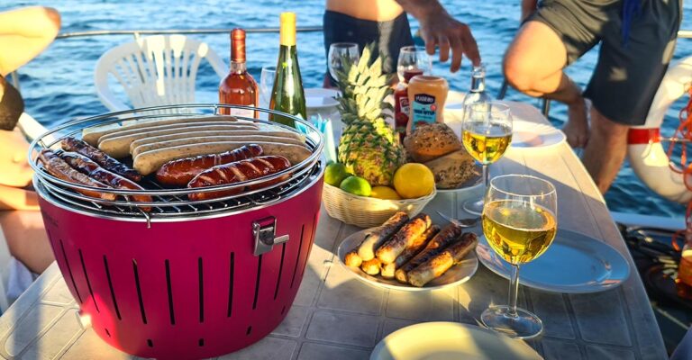 Ein gedeckter Tisch mit einem Grill auf dem bereits einige Würstchen liegen. Daneben ist ein Teller mit bereits Fertigen Würstchen. Dazu ein Paar Gläser Wein, ein Korb mit Obst und ein Paar Brötchen.