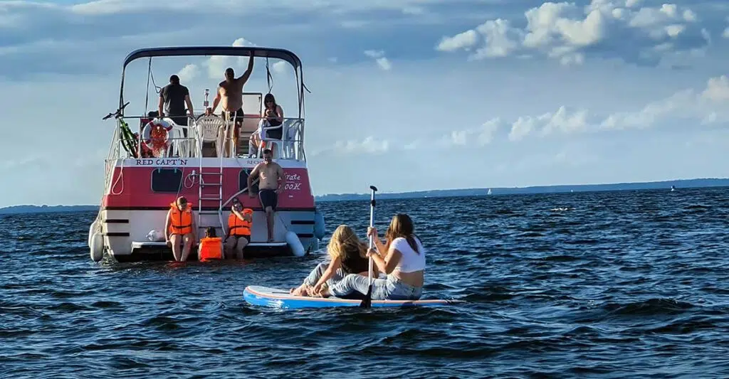 Zwei Mädchen auf einem SUP Paddeln zur Pirate. Auf der Badeplattform sind drei Personen. Eine Person ist im Wasser. Drei Personen befinden sich auf dem Sonnendeck.