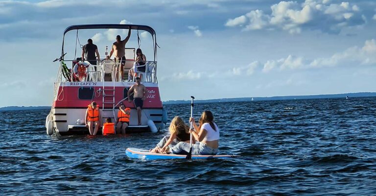 Zwei Mädchen auf einem SUP Paddeln zur Pirate. Auf der Badeplattform sind drei Personen. Eine Person ist im Wasser. Drei Personen befinden sich auf dem Sonnendeck.