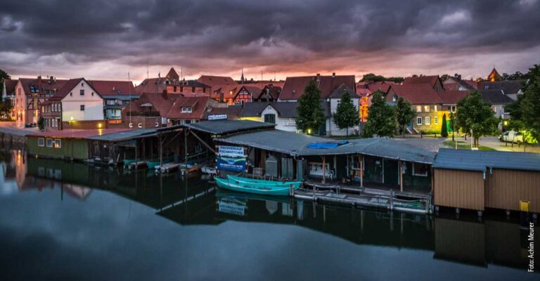 Luftaufnahme mit einer Reihe Bootshäuser, u.A das der Müritzfischer. im Hintergrund ist die Stadt Plau am See zu sehen.