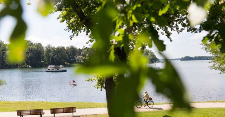 Ein Radweg der am Wasser entlang führt, zwei Radfahrer fahren gerade entlang.