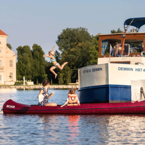 Ein Kanu vor einer Kormoran mit zwei Personen. Auf der Kormoran sind drei Personen. Ein Kind springt ins Wasser.