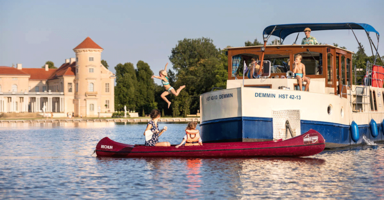 Ein Kanu vor einer Kormoran mit zwei Personen. Auf der Kormoran sind drei Personen. Ein Kind springt ins Wasser.