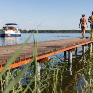 Zwei Personen auf einem Steg gehen in Richtung Wasser. Am ende des Steges springt bereits eine weiter Person ins Wasser