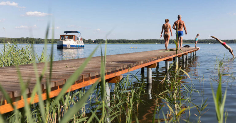 Zwei Personen auf einem Steg gehen in Richtung Wasser. Am ende des Steges springt bereits eine weiter Person ins Wasser