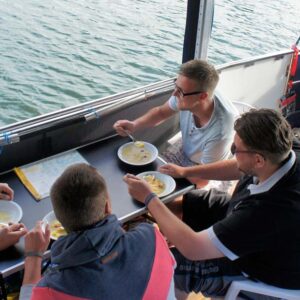 Eine Gruppe sitzt an einem Campingtisch auf dem Febo720 open und isst Suppe.