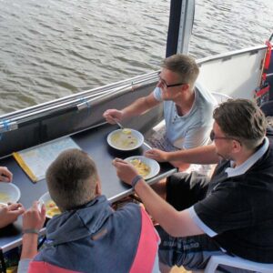 Eine Gruppe sitzt an einem Campingtisch auf dem Febo720 open und isst Suppe.