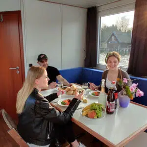 Drei Personen sitzen im Salon des Saunahausbootes. Auf dem Tisch stehen drei Gläser mit Weißwein, drei Flaschen sowie Obst und Brötchen.