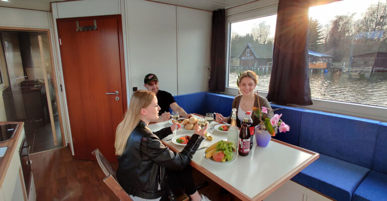 Drei Personen sitzen im Salon des Saunahausbootes. Auf dem Tisch stehen drei Gläser mit Weißwein, drei Flaschen sowie Obst und Brötchen.