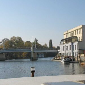 Eine Kormoran auf einem Kanal in Brandenburg mit Blick auf ein Restaurant und einer Brücke.