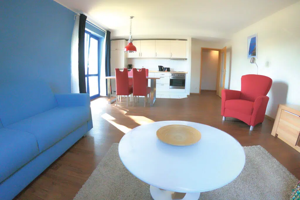 Wohnzimmer mit Couch und Sessel. Blick ist auf die Kuchenzeile, den Esstisch und auf die Tür zum Balkon.