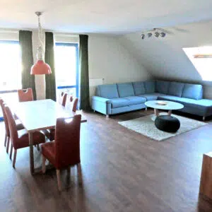Wohnzimmer mit großer Eckcouch, Sessel, Fernseher, großem Esstisch mit sechs plätzen und Tür zum Balkon.