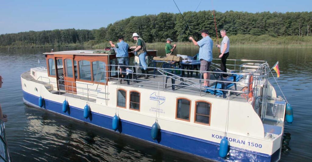 Das Hausboot Kormoran auf einem See mit 5 Anglern an Deck