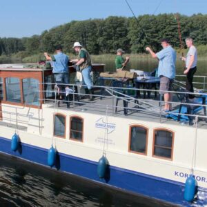 Das Hausboot Kormoran auf einem See mit 5 Anglern an Deck