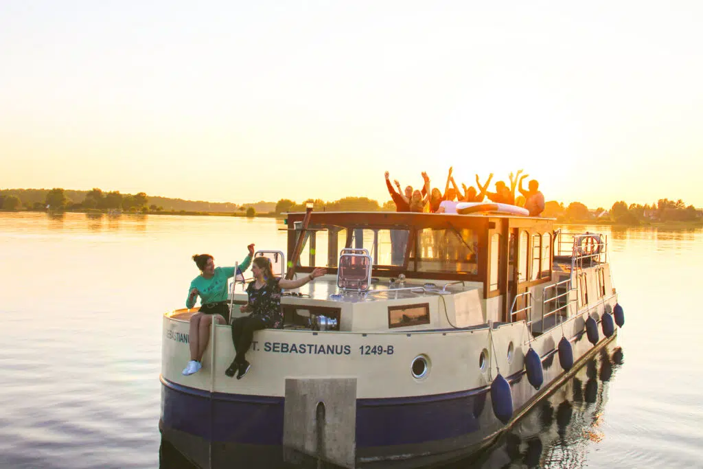 Kormoran 1500 fährt mit einer Gruppe von Reisenden auf dem Deck, sie fahren auf einem See entgegen des Sonnenuntergangs