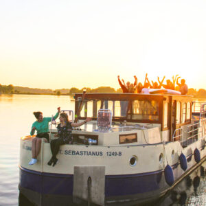 Kormoran 1500 fährt mit einer Gruppe von Reisenden auf dem Deck, sie fahren auf einem See entgegen des Sonnenuntergangs