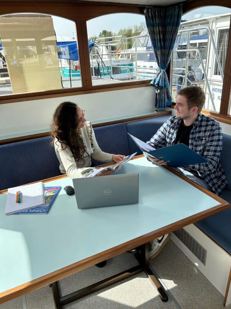 Zwei junge Personen sitzen sich in einem Boot gegenüber. Ein mann und eine Frau sprechen miteinadner über die Arbeit, er hat eine Akte in der Hand und sie analysiert einen Bericht.