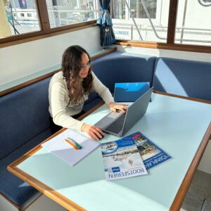 Junge Frau sitzt auf einem Boot mit einem Laptop vor sich und arbeitet. Auf dem Tisch vor ihr liegen verschiedene Prospekte und Ordner. Draußen scheint die Sonne.