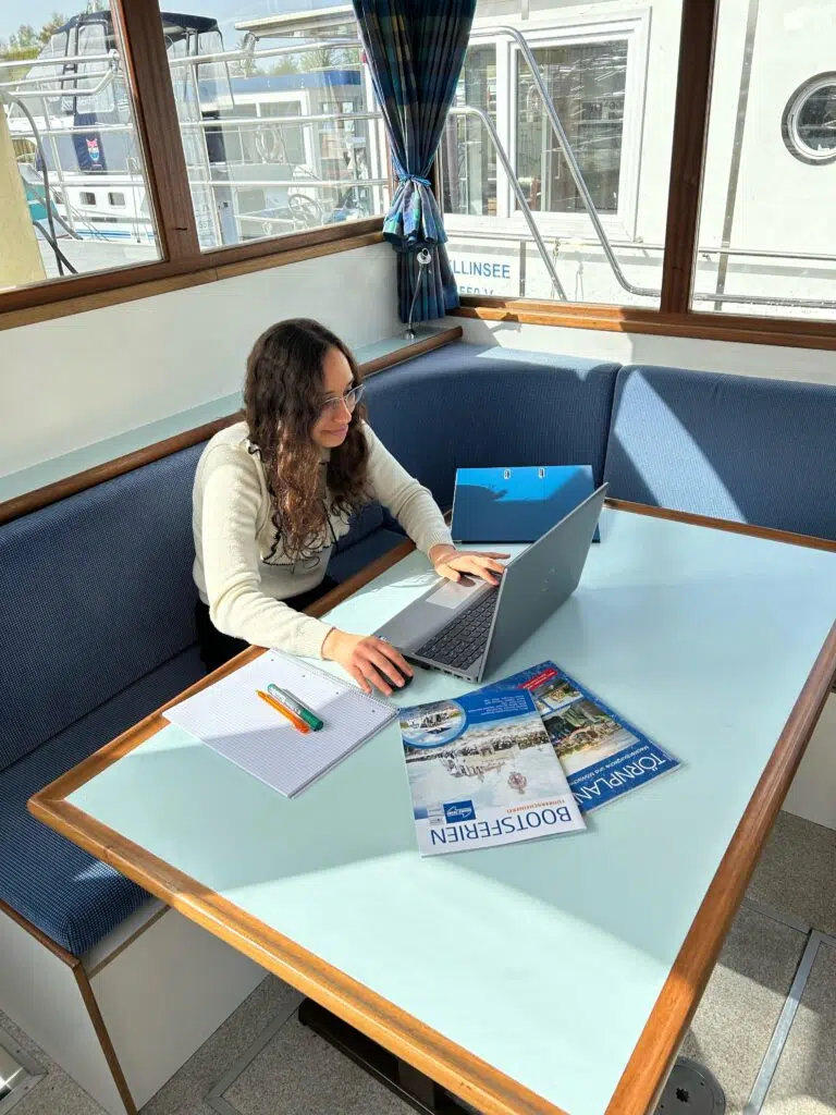 Junge Frau sitzt auf einem Boot mit einem Laptop vor sich und arbeitet. Auf dem Tisch vor ihr liegen verschiedene Prospekte und Ordner. Draußen scheint die Sonne.